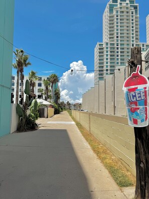 A sidewalk with a trash can, a palm tree, and a tall building in the background. - 6 Beds | Pool Steps Away | Private Beach Access (South Padre Island)