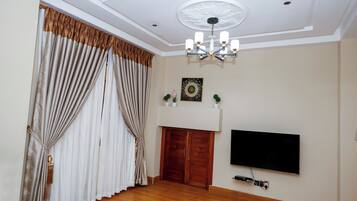 Une pièce dotée d’un plancher en bois, d’un téléviseur à écran plat fixé au mur, d’un plafond agrémenté d’un luminaire décoratif et d’un ensemble de rideaux.