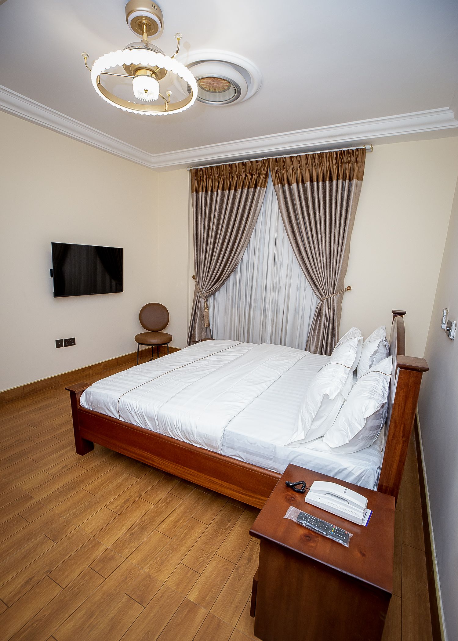 Une chambre avec un lit en bois, une table de chevet avec un téléphone et une télécommande, un téléviseur à écran plat fixé au mur et un luminaire au plafond.