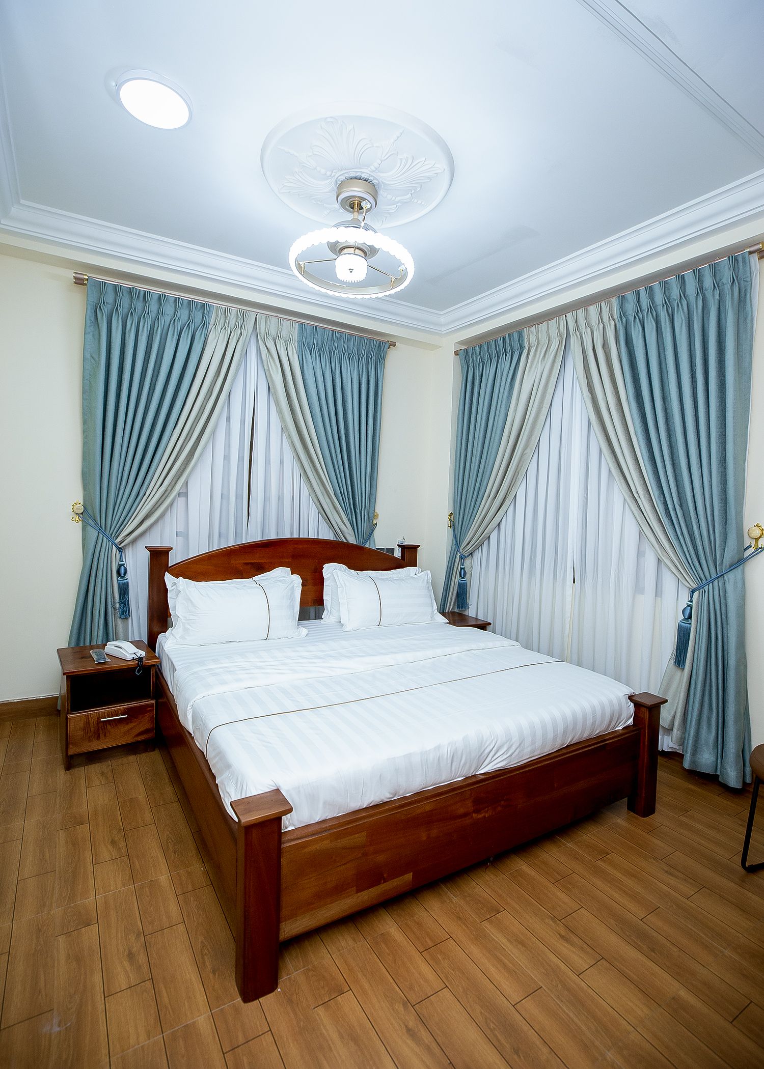Ein ordentlich bezogenes Bett mit Holzbettgestell und weißer Bettwäsche, ein Nachttisch mit Lampe und ein Deckenventilator.