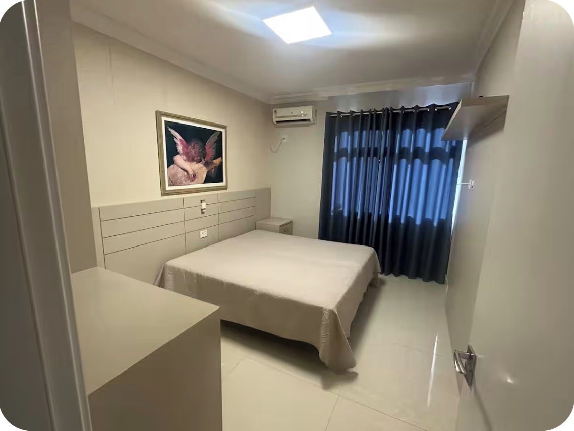 Quarto de hotel com cama, mesa de cabeceira, pequena escrivaninha, quadro na parede e janela com cortinas.