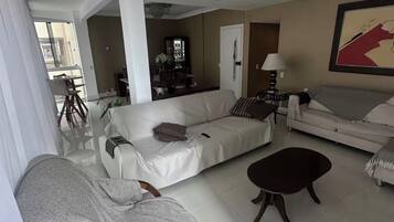 Uma sala de estar ampla com sofá, poltrona, mesa de centro e uma área de jantar ao fundo.
