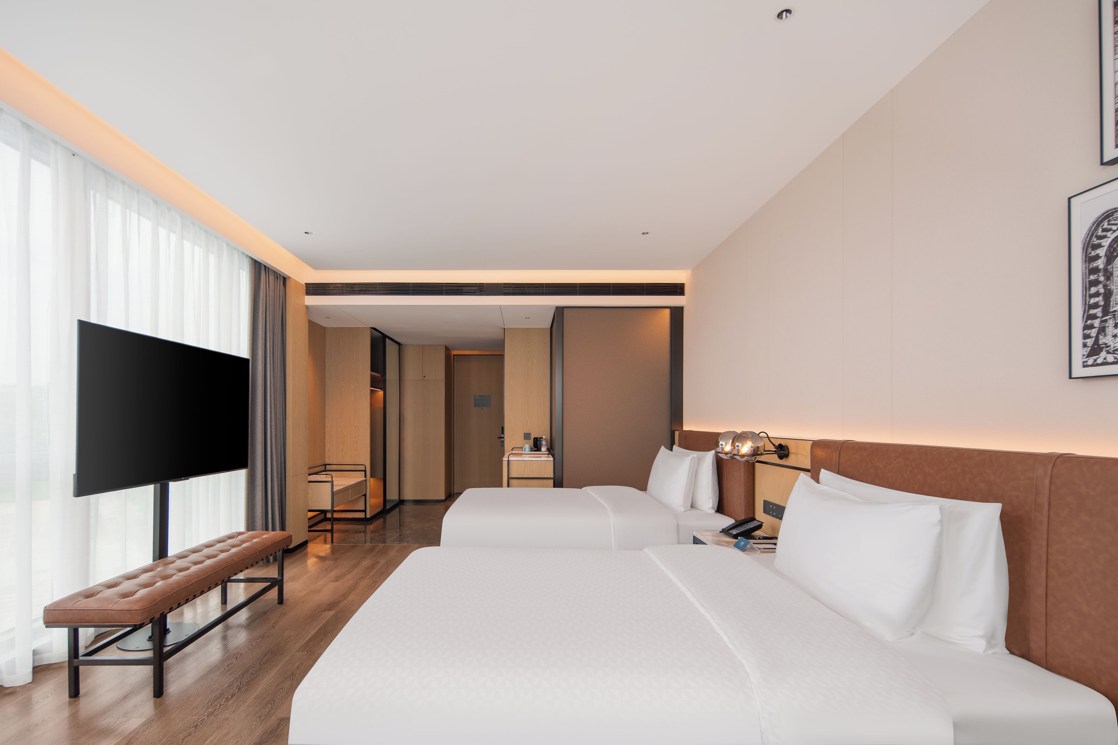 A hotel room with a large flat-screen TV, a bench, two beds, a bedside table with a lamp, and a framed picture on the wall.