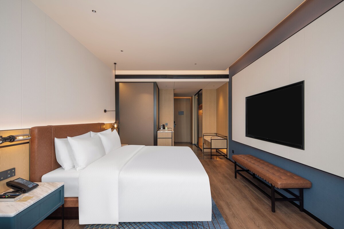 A hotel room with a bed, bedside table, lamp, TV, and a bench.
