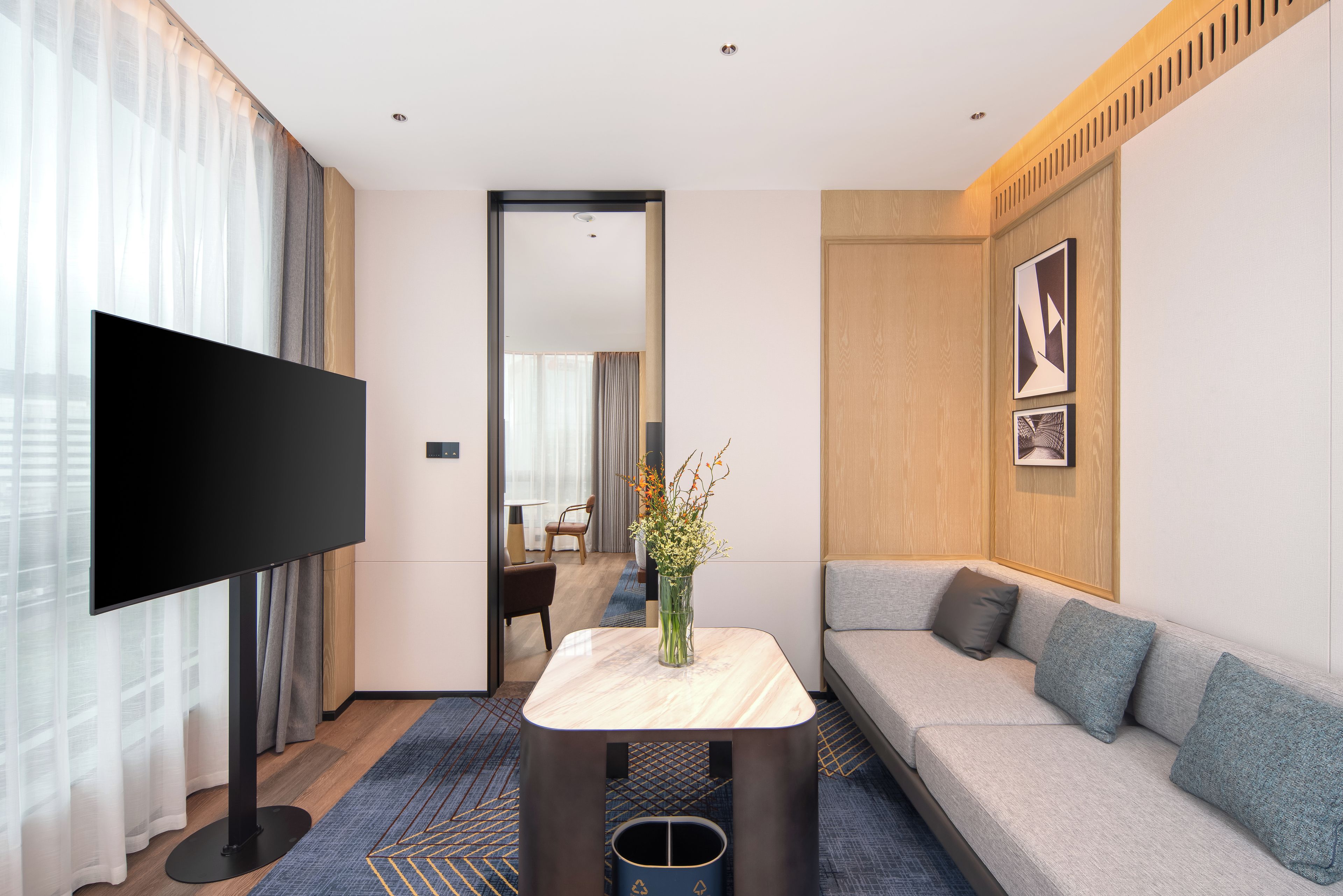 A modern hotel room with a flat-screen TV, a sofa, a small table with a vase, and a mirror reflecting the room.