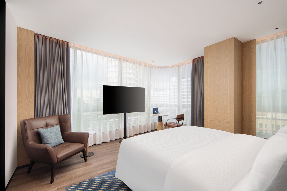 A hotel room with a large bed, a flat-screen TV mounted on the wall, a seating area with a chair and a small table, and large windows with sheer curtains.