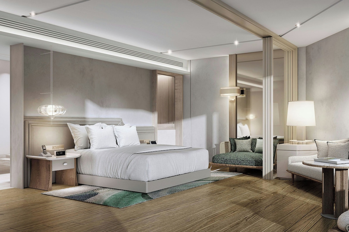 A modern hotel room with a large bed, bedside tables, a seating area, and a mirror.