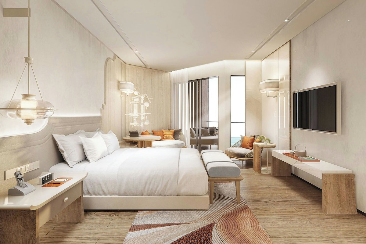 A modern hotel room with a large bed, bedside tables, a TV, and a view of the sea.