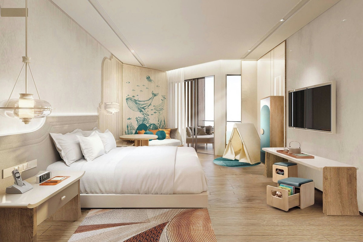 A modern hotel room with a large bed, bedside tables, a desk, a chair, a TV, and a wall mural.