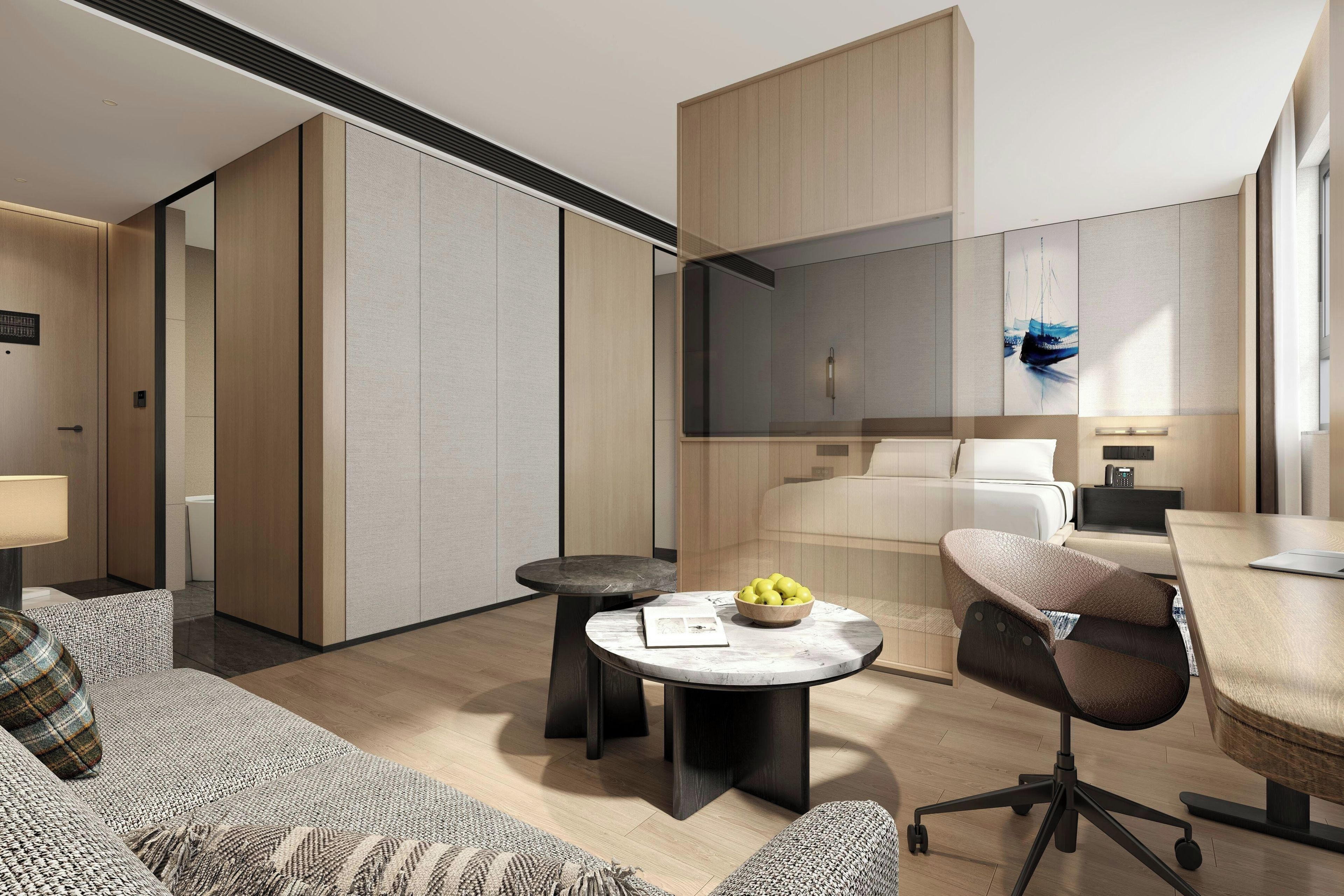 Una habitación de hotel moderna con sofá, una mesa redonda con frutero, un escritorio con computadora portátil y cama con cabecera blanca.