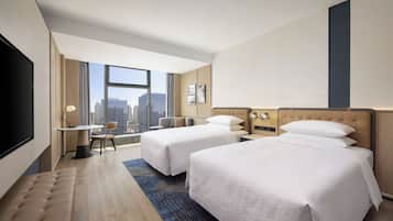 A hotel room with two beds, a desk, and a large window offering a city view.