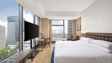 Una habitación de hotel moderna con una cama grande, un escritorio, una silla, un televisor y vistas a la ciudad.