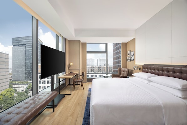A modern hotel room with a large bed, a desk, a chair, a television, and a view of the cityscape.