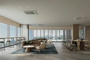Un amplio vestíbulo de hotel con techo alto, ventanas grandes que ofrecen vista a la ciudad, una zona de estar con una mesa redonda y un área de bar.