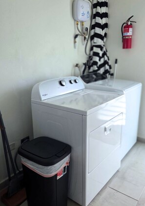 A front-loading washing machine with a control panel, a black trash bin, and a fire extinguisher mounted on the wall. - Azulena - A stay between the mountains & sea! With Tesla battery & water tank (Fajardo)