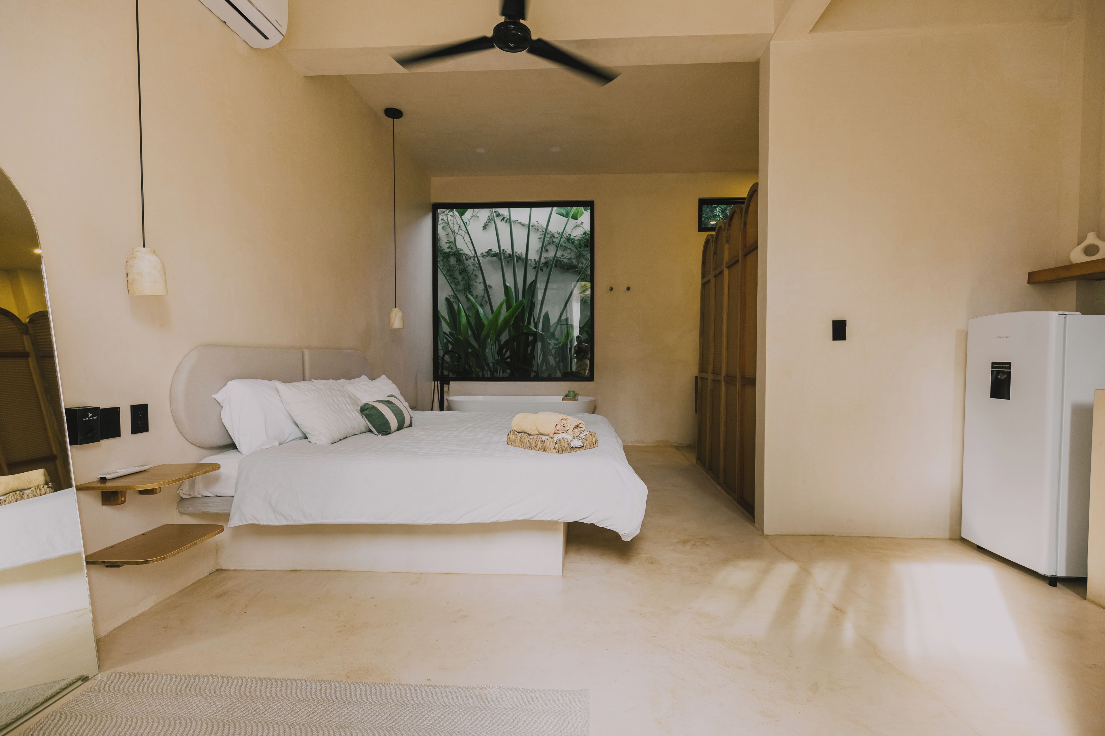 Un dormitorio con cama, ventilador de techo, ventana con vistas a la vegetación y un refrigerador blanco.
