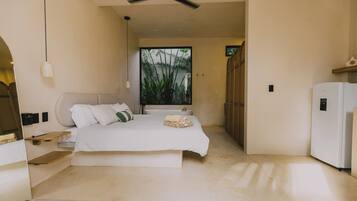 Un dormitorio con cama, ventilador de techo, ventana con vistas a la vegetación y un refrigerador blanco.