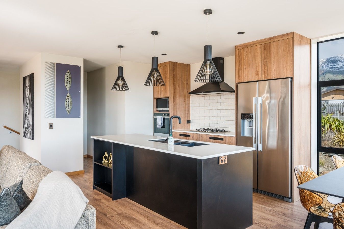 A modern kitchen with a central island, stainless steel appliances, pendant lighting, and a view through a window.