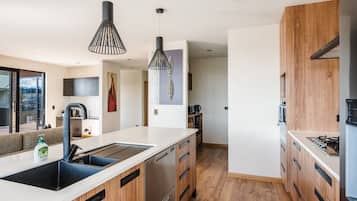 A modern kitchen with a central island, wooden cabinets, stainless steel appliances, pendant lighting, and a view to the outside.