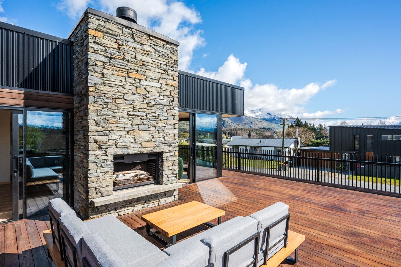 A modern outdoor patio with a stone fireplace, wooden furniture, and a view of mountains.