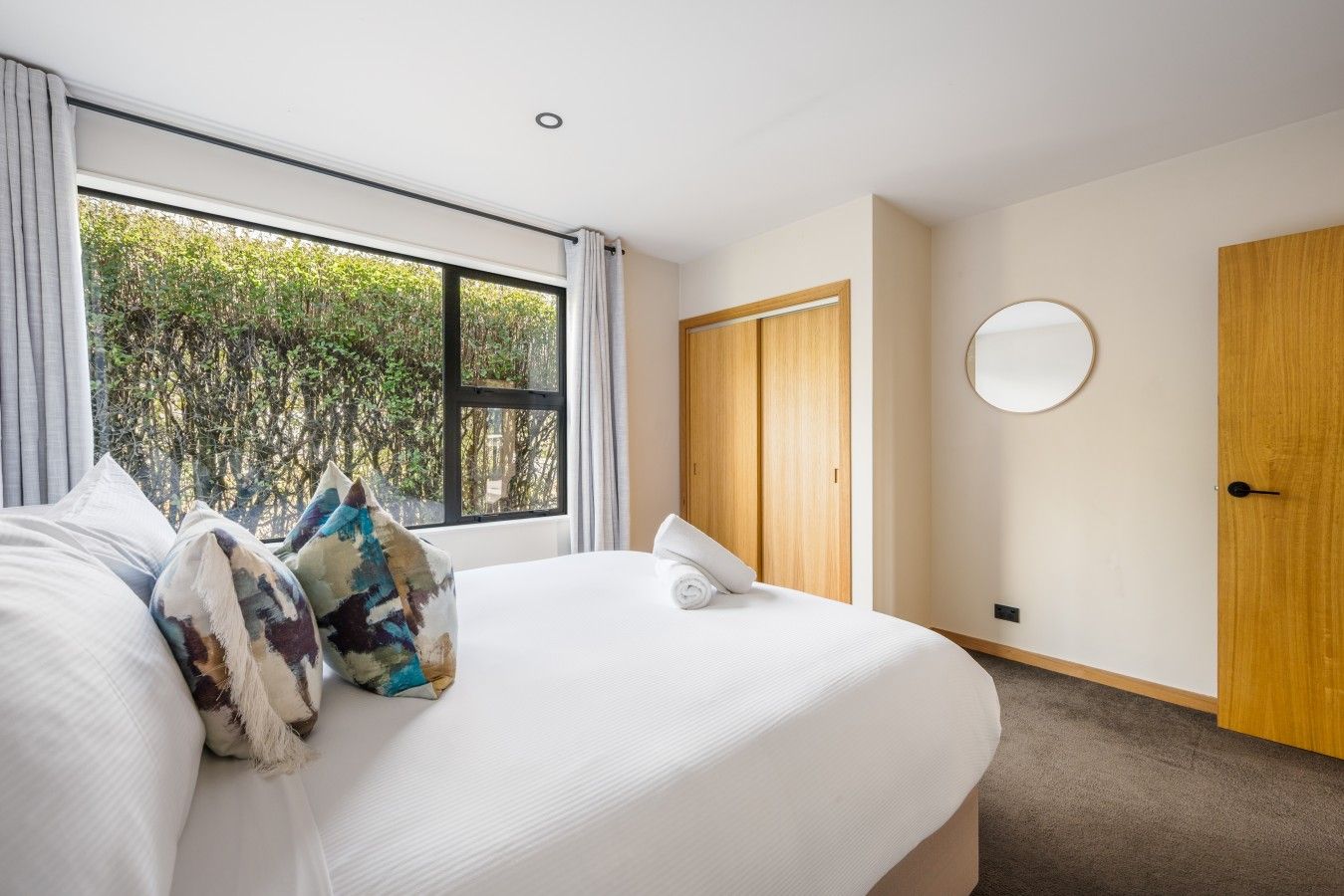 Ein Hotelzimmer mit einem großen Bett, einem Holzschrank, einem runden Spiegel und einem Fenster mit Blick auf Grünflächen.