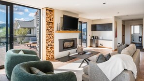 Een moderne woonkamer met een open haard, een flatscreen televisie en een balkon met uitzicht op een huis en bomen.
