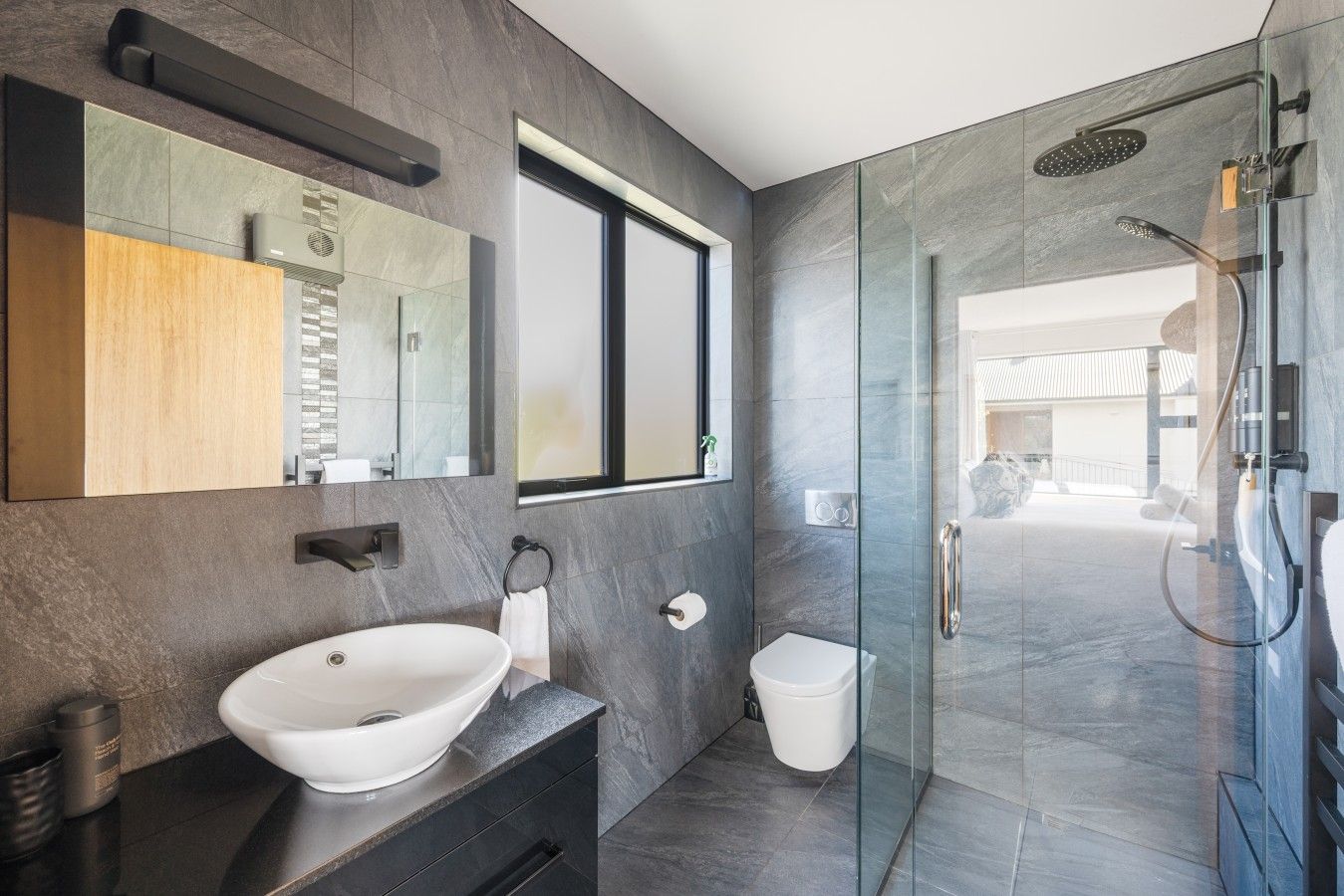 A modern bathroom with a large mirror, a vessel sink, a glass-enclosed shower, and a view of the outdoor area.
