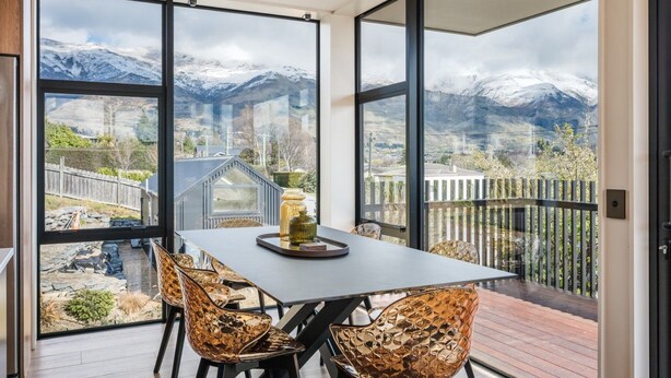 Een eetruimte met een tafel en stoelen, grote ramen met uitzicht op de bergen, en een houten terras buiten.