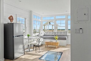 A modern kitchen with a refrigerator, dining table, and chairs. There is a seating area with a sofa and a window overlooking a beach. - Ocean Front Boardwalk Nest with Direct Beach Access (Santa Monica)