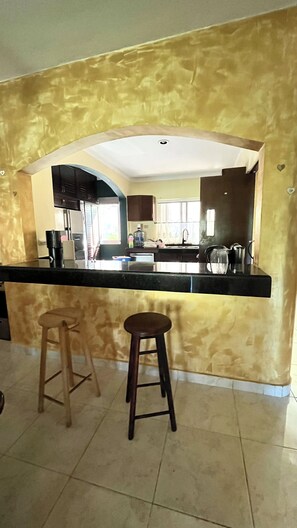 A kitchen with a black countertop, wooden stools, and a view of the adjacent dining area.