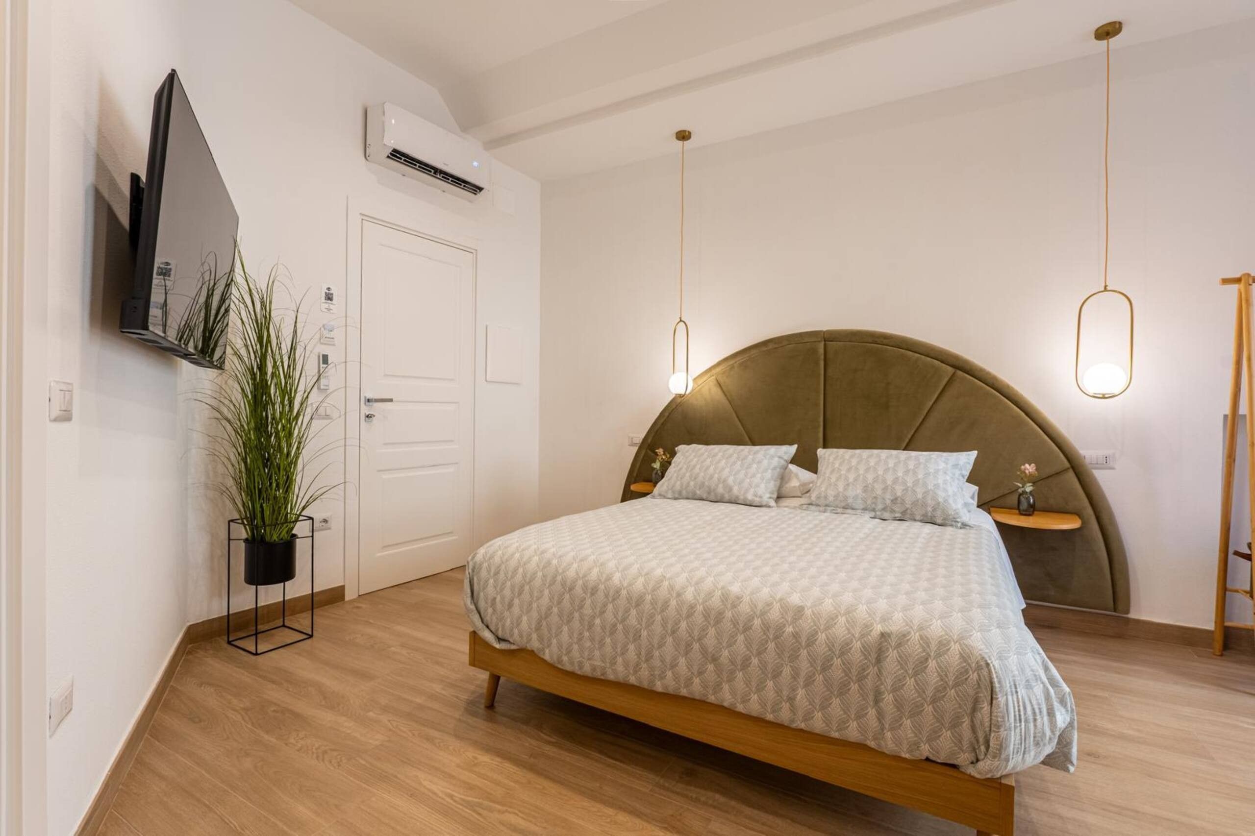 A bedroom with a bed, a television mounted on the wall, an air conditioning unit, and a potted plant.