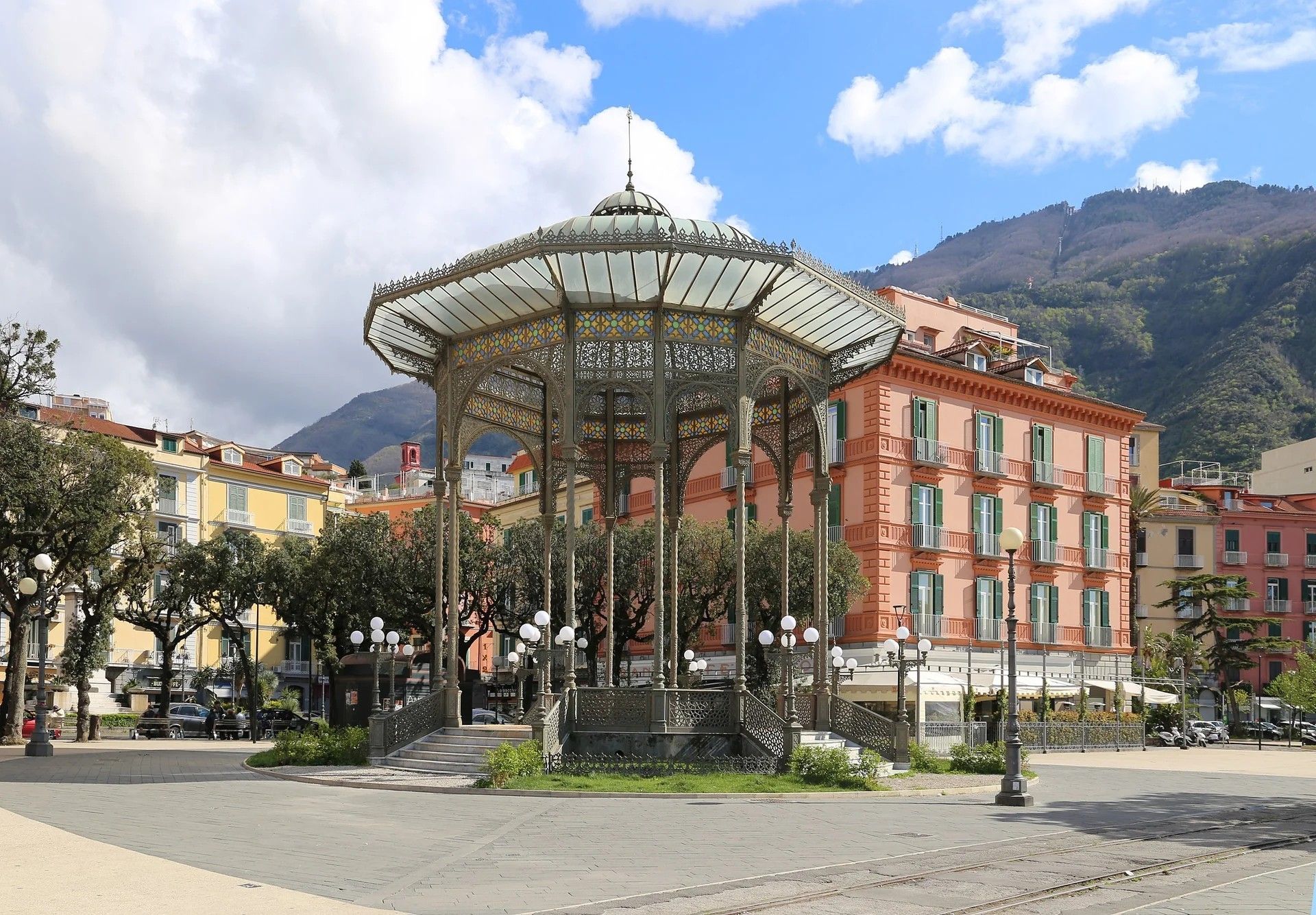A gazebo with a dome and ornate metalwork, surrounded by a paved plaza and buildings with balconies.