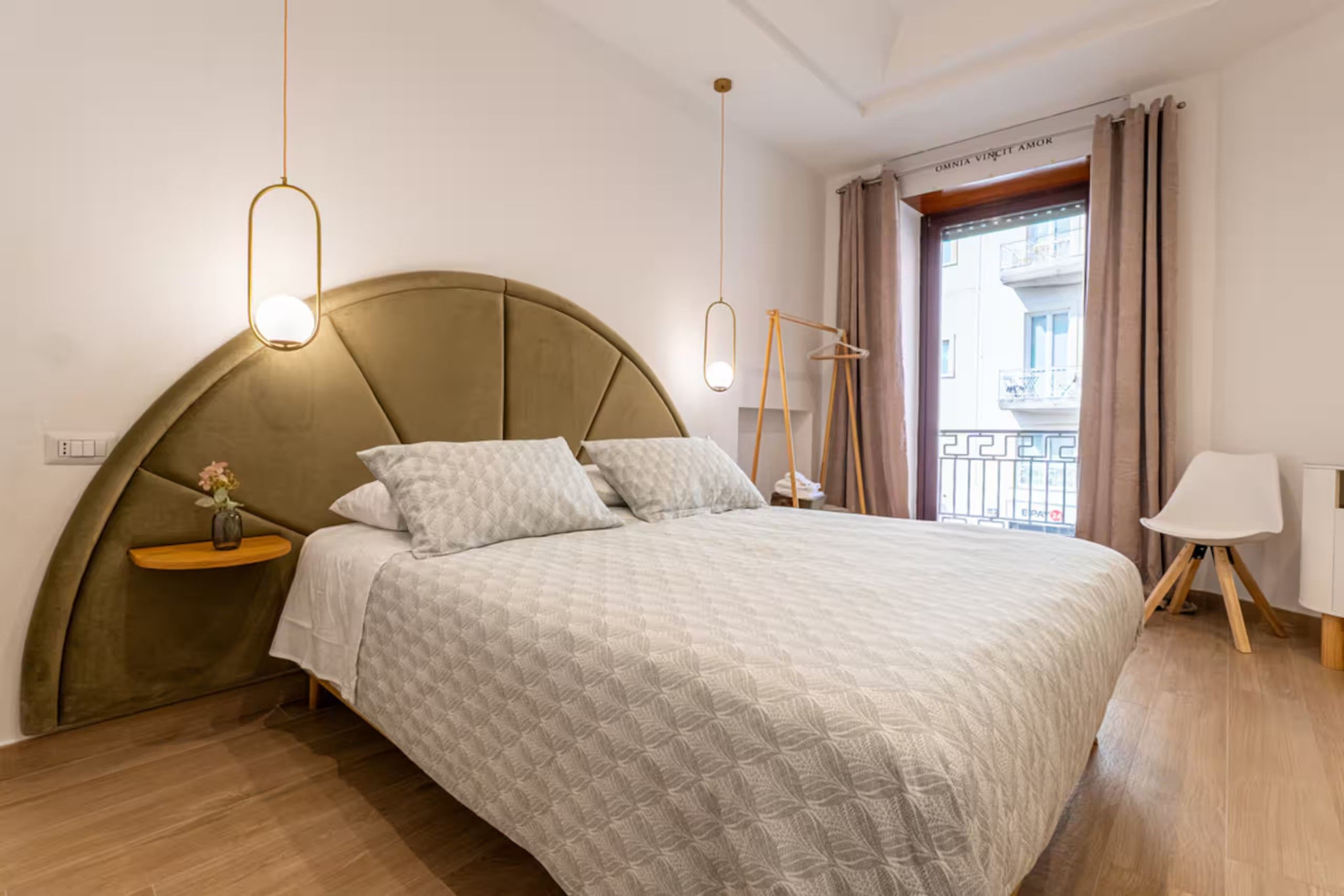 A modern bedroom with a curved headboard, a bed with a patterned duvet, bedside tables, a hanging light fixture, and a view of the outside through a window with curtains.