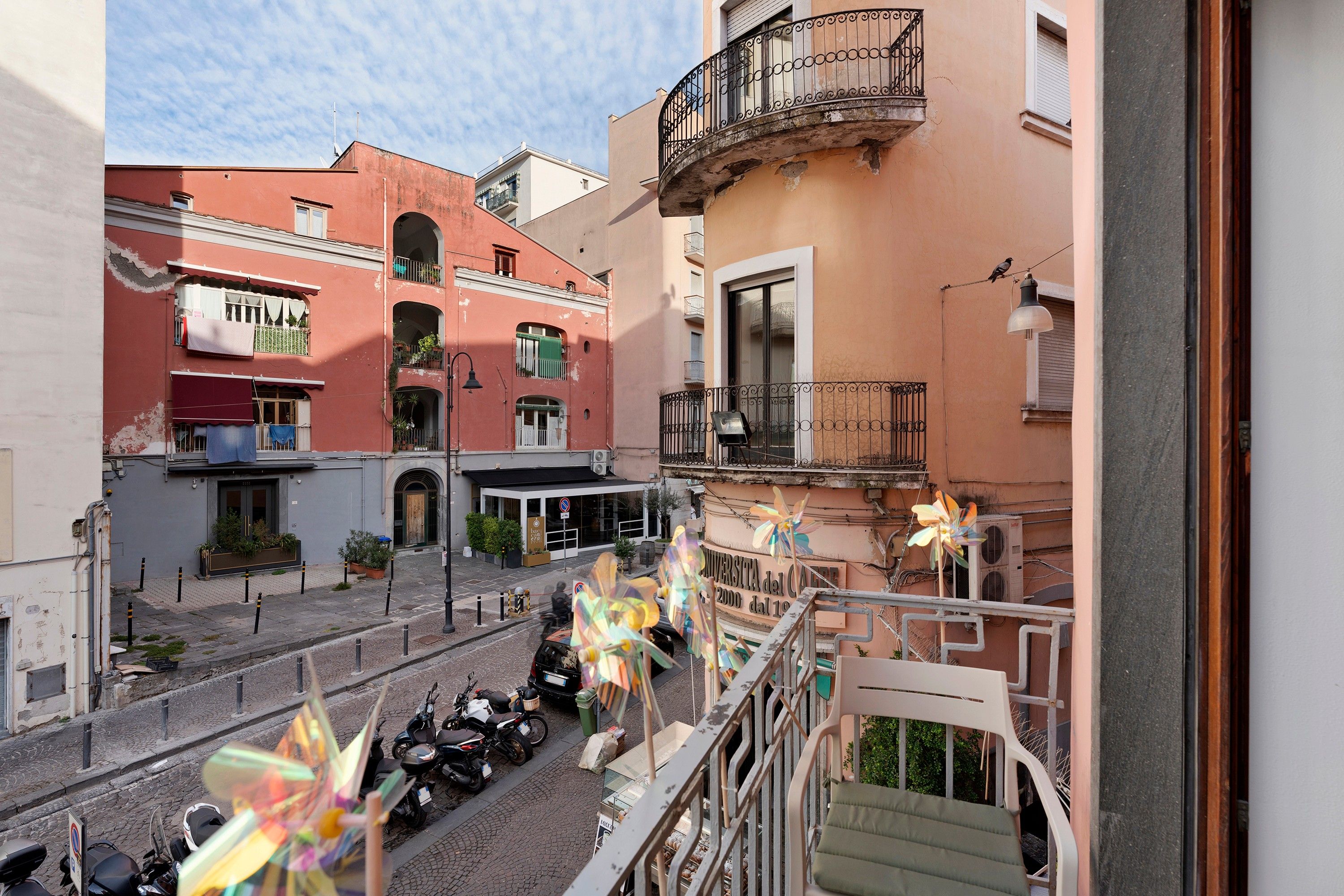 Un cortile con scooter parcheggiati, un edificio con un balcone e un cartello con la scritta 'Università del Cale'.