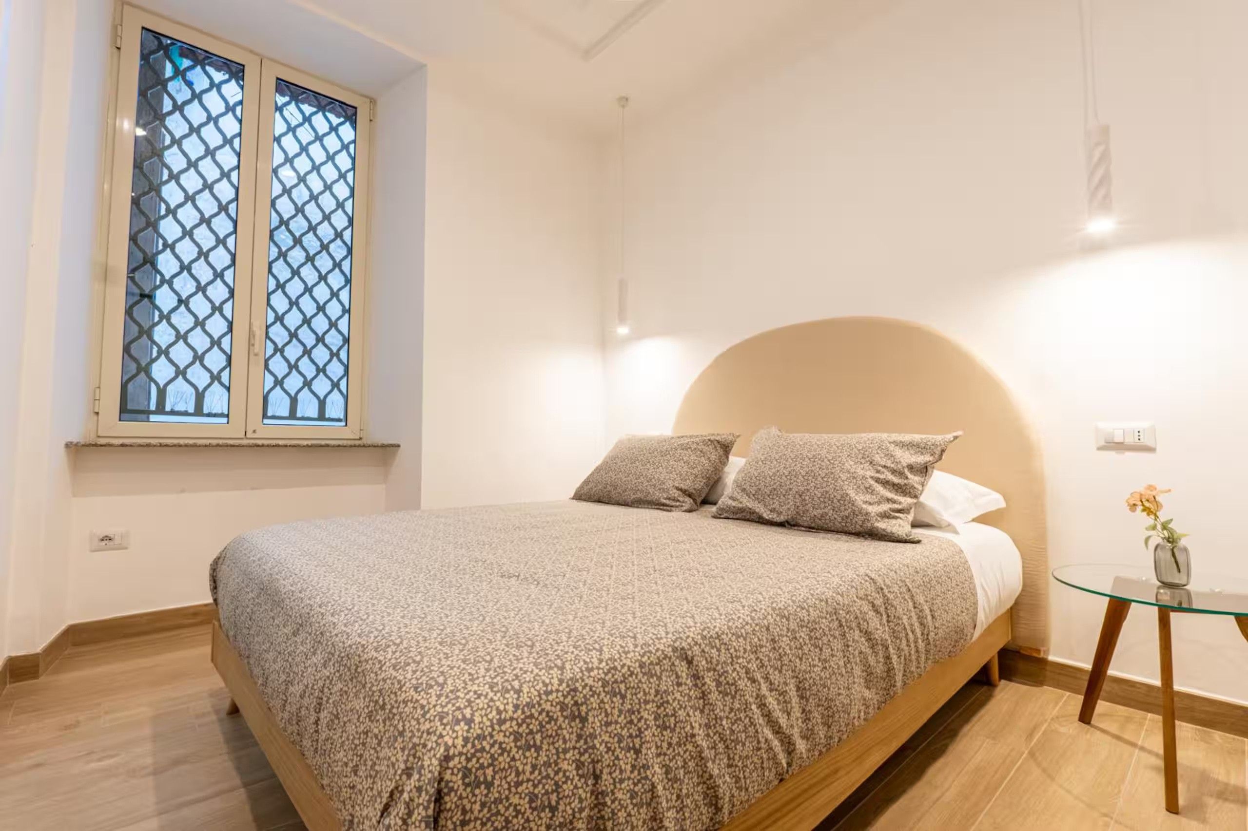 Ein Schlafzimmer mit einem Bett, einem Fenster mit Insektenschutzgitter, einem Nachttisch mit einer Vase und einer Steckdose.