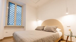 Ein Schlafzimmer mit einem Bett, einem Fenster mit Insektenschutzgitter, einem Nachttisch mit einer Vase und einer Steckdose.
