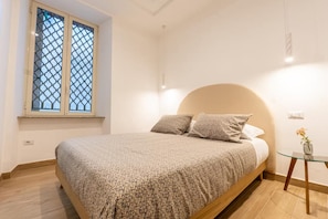 Een slaapkamer met een bed, een raam met hor, een nachtkastje met een vaas en een stopcontact.