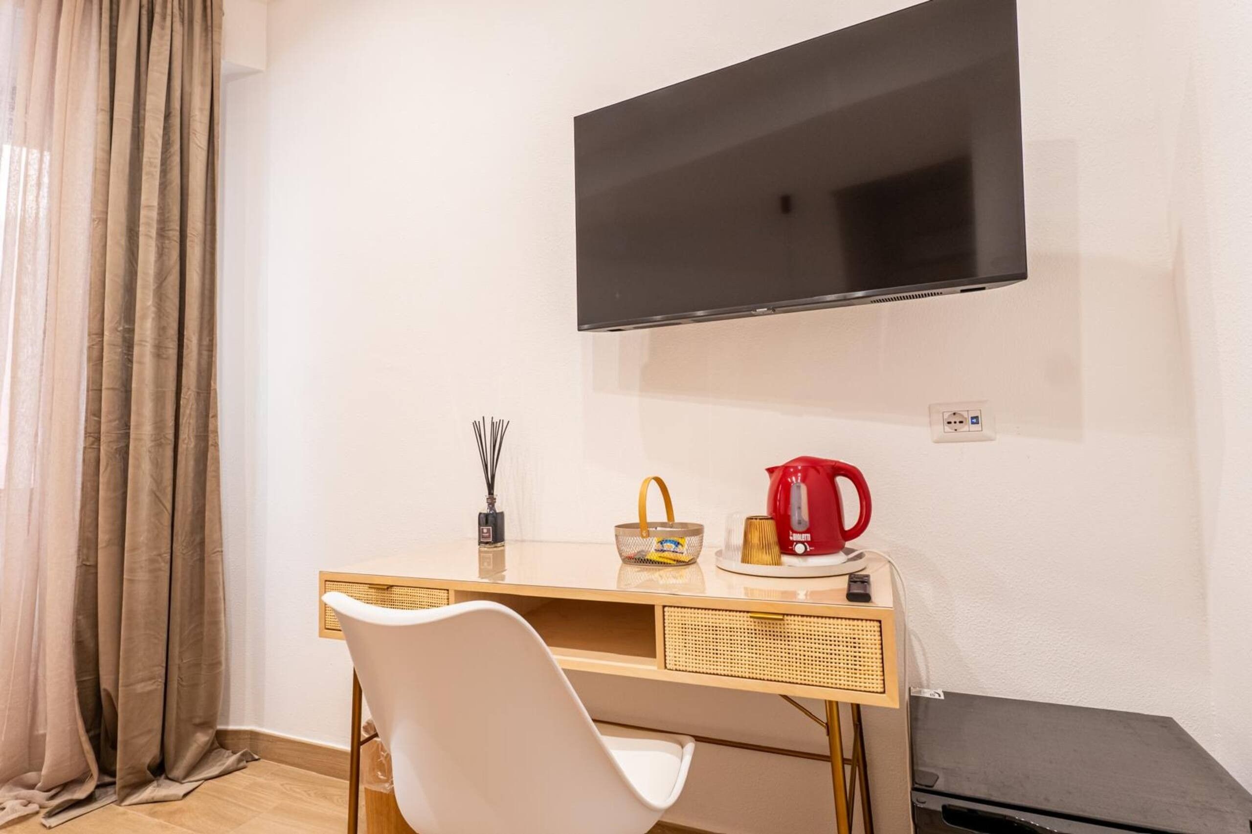 Una habitación con televisión de pantalla plana en la pared, un escritorio de madera con silla, una tetera roja y una cesta pequeña.