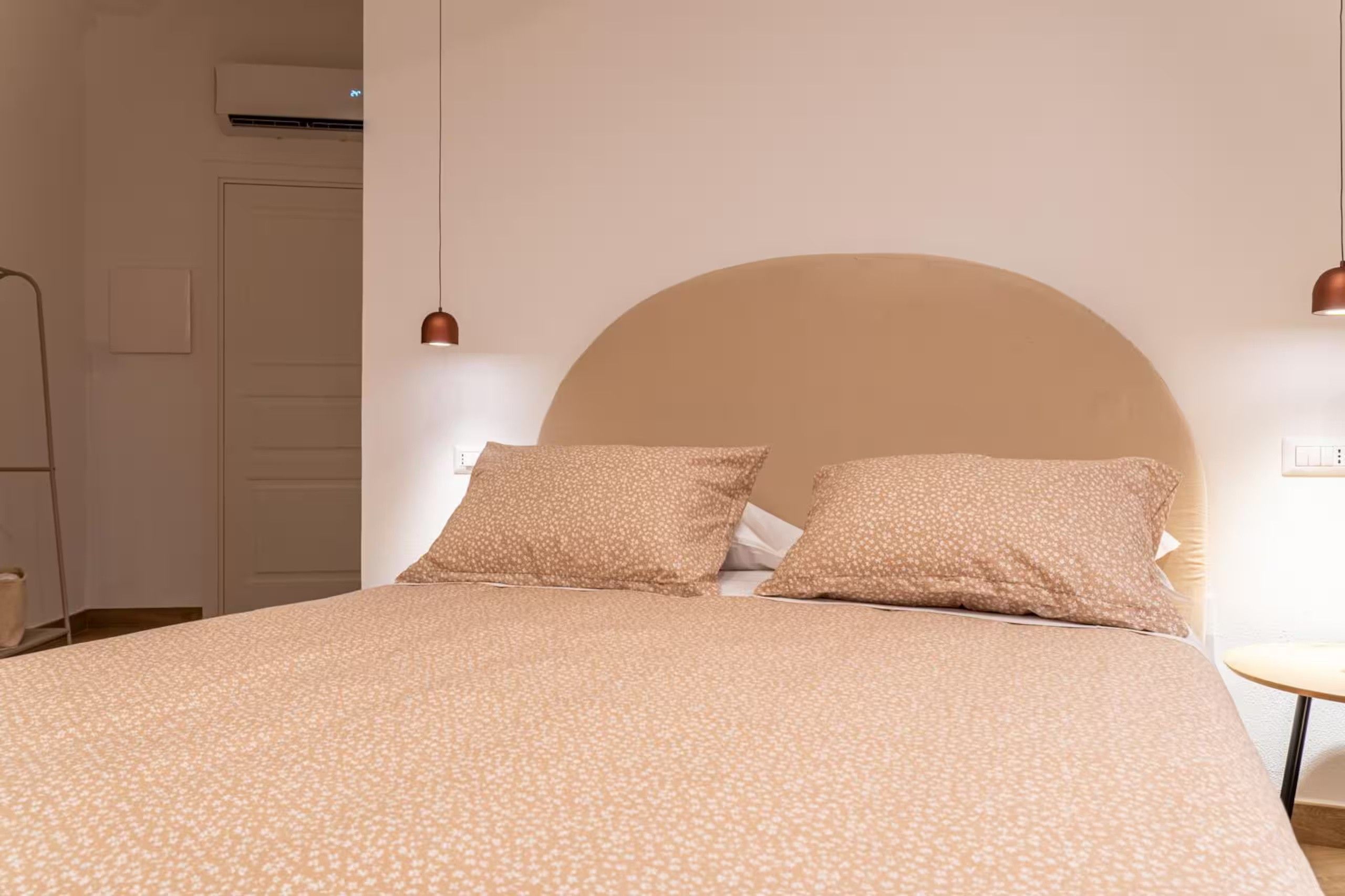 A bed with a large headboard, two pillows, and a patterned bedspread in a room with a white wall and a small table.