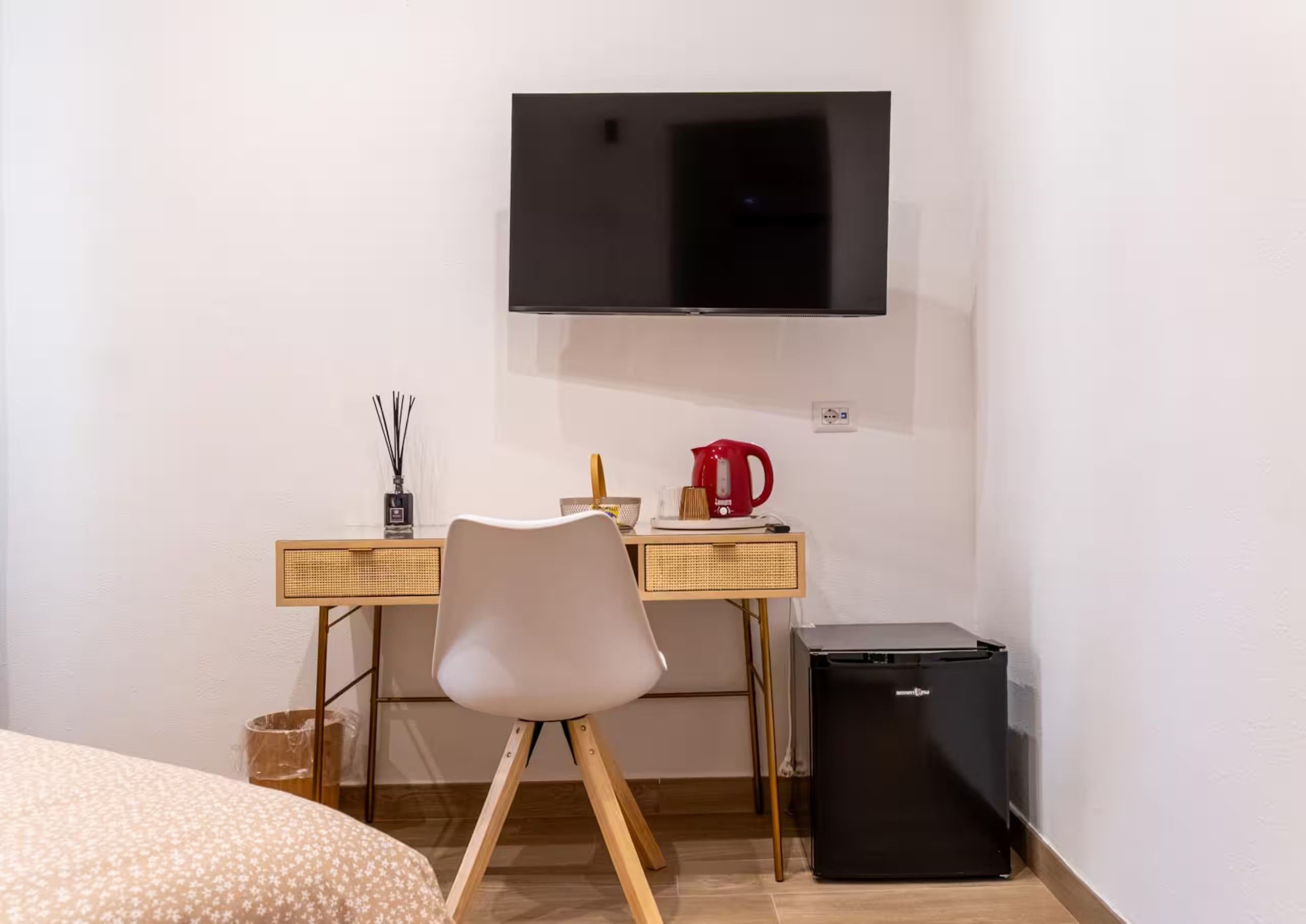 A room with a bed, a desk with a chair, a television mounted on the wall, a red kettle, and a small black refrigerator.