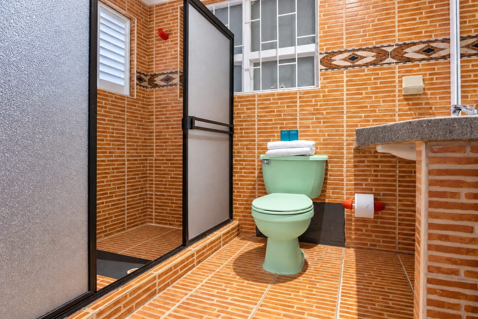 Un baño con inodoro verde, zona de ducha con puerta de cristal y suelo de baldosas.