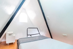 A small, triangular room with a bed, a chair, and a white nightstand. - Hotel Bogota Home (Bogotá)