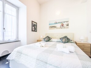 A neatly made bed with two pillows, a window with blinds, a painting of a bicycle on a bridge, and a bedside lamp. - Marienkäfer by Interhome (Corniglia)