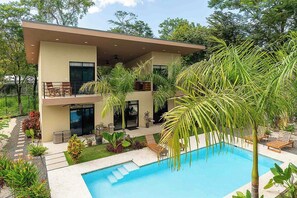 Et moderne hus i to etager med swimmingpool, udendørs møbler og frodig beplantning.