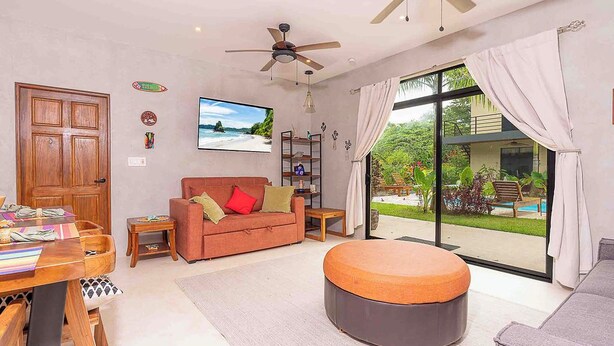 Ein geräumiges Wohnzimmer mit großem Fenster, Deckenventilator, einer Couch, einem Couchtisch und einem Essbereich mit Holztisch und Stühlen.