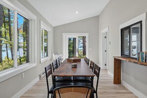 Un comedor amplio con una larga mesa de madera, varias sillas y vistas a árboles a través de ventanas grandes.