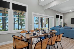 Un coin repas avec une table en bois et des chaises en osier, de grandes fenêtres offrant une vue sur les arbres et l’eau, et un canapé bleu en arrière-plan.