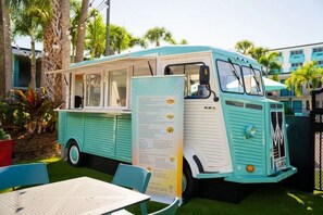 Ein Vintage-Foodtruck steht draußen, mit einer Speisekarte, umgeben von Palmen und einem Gebäude im Hintergrund.