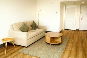 Ein Zimmer mit einem beigefarbenen Sofa, zwei Kissen, einem kleinen Holfnachtkästchen, einem runden Couchtisch und einem gewebten Teppich auf Holzböden.