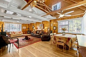 A spacious living room with a fireplace, wooden flooring, a dining area, and large windows. - Red Door Cottage. Close to town! Pet-friendly! (Twain Harte)